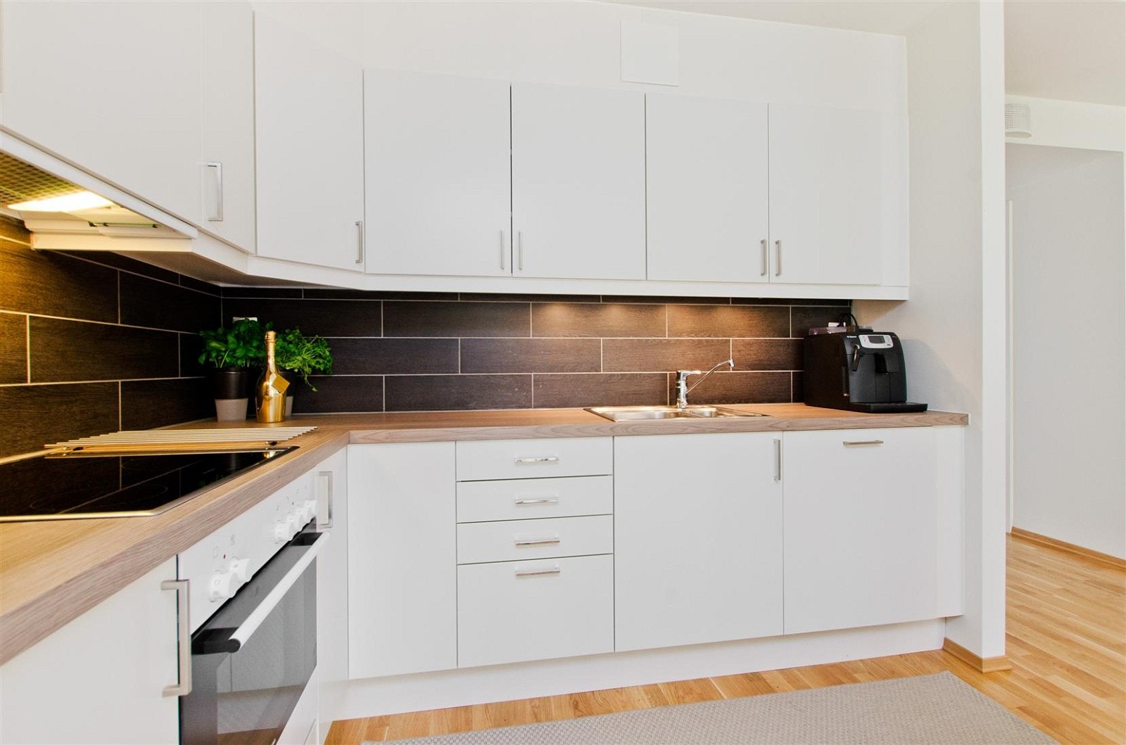 Hvite glatte fronter, takhøye overskap med lys under, flisfelt over kjøkkenbenk og nedfelt kjøkkenbeslag. Galleribilde