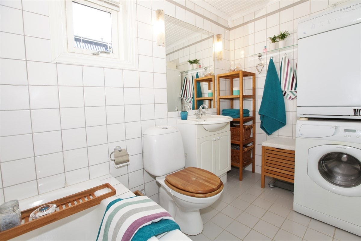 Flislagt baderom med varmekabler i gulv, badekar m/ dusjvegg i glass, toalett, servant m/ underskap og opplegg for vaskesøyle Galleribilde