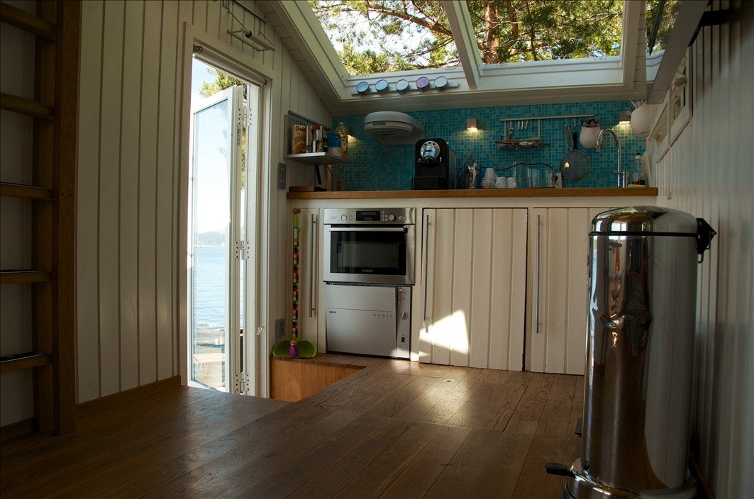 Kjøkken med komfyr, induksjonsplater, avtrekksvifte, oppvaskmaskin, kjøleskap, fryseboks, isbitmaskin, skarpe kniver og åpent tak til furutoppen utenfor. Galleribilde