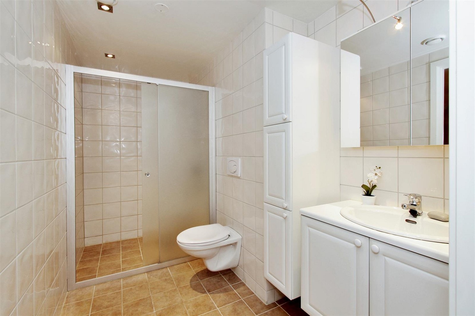 Badet har hvit profilert innredning, speilskap, veggheng wc og stor dusjnisje Galleribilde