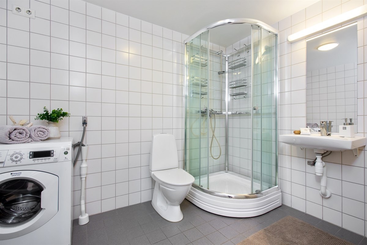 Det er satt opp et praktisk dusjkabinett i hjørnet, og det er opplegg til vaskemaskin og tørketrommel langs veggen. Servantskap i hvit utførelse med speil. Galleribilde