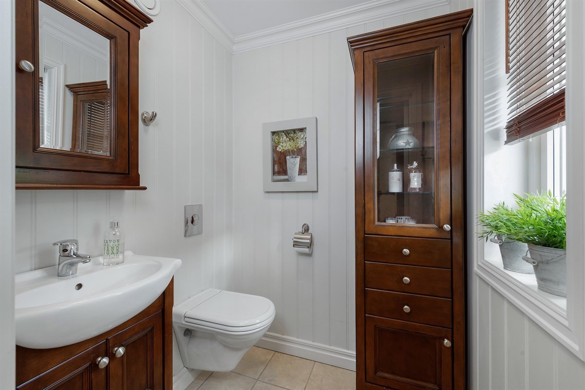 Separat wc har lyse fliser på gulv, hvitmalte vegger, enkel servant på gyllenbrunt servantskap, matchende høyskap og speilskap, samt veggmontert toalett, og et vindu som gir naturlig lys. Galleribilde