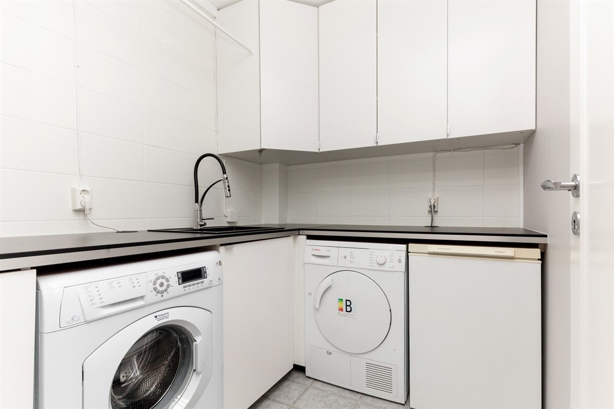 Nyoppusset og praktisk innredet vaskerom med arbeidsbenk med utslagsvask, overskap og opplegg for vaskemaskin og tørketrommel Galleribilde