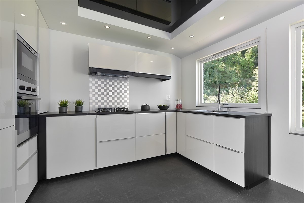 Innredningen har hvite, høyglansfronter med mørk omramming og benkeplate i mørk laminat med nedfelt vaskekum. Det er lekker stålplate og vindu over benk. Galleribilde