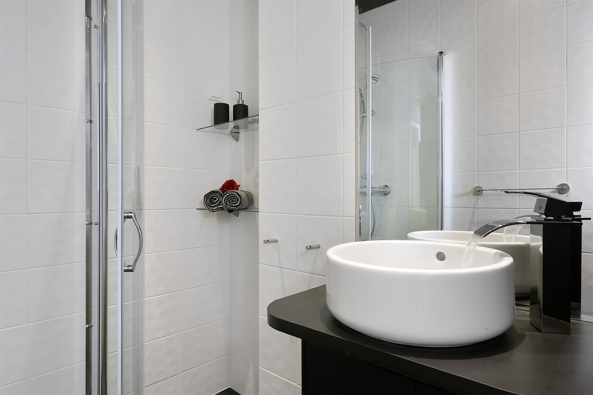 Rommet har rund porselensservant med designarmatur, mørkt servantskap, matchende høyskap og speil. Videre er det veggmontert toalett, samt et flott dusjhjørne med innfellbare glassdører og regnfallsdusj. Galleribilde