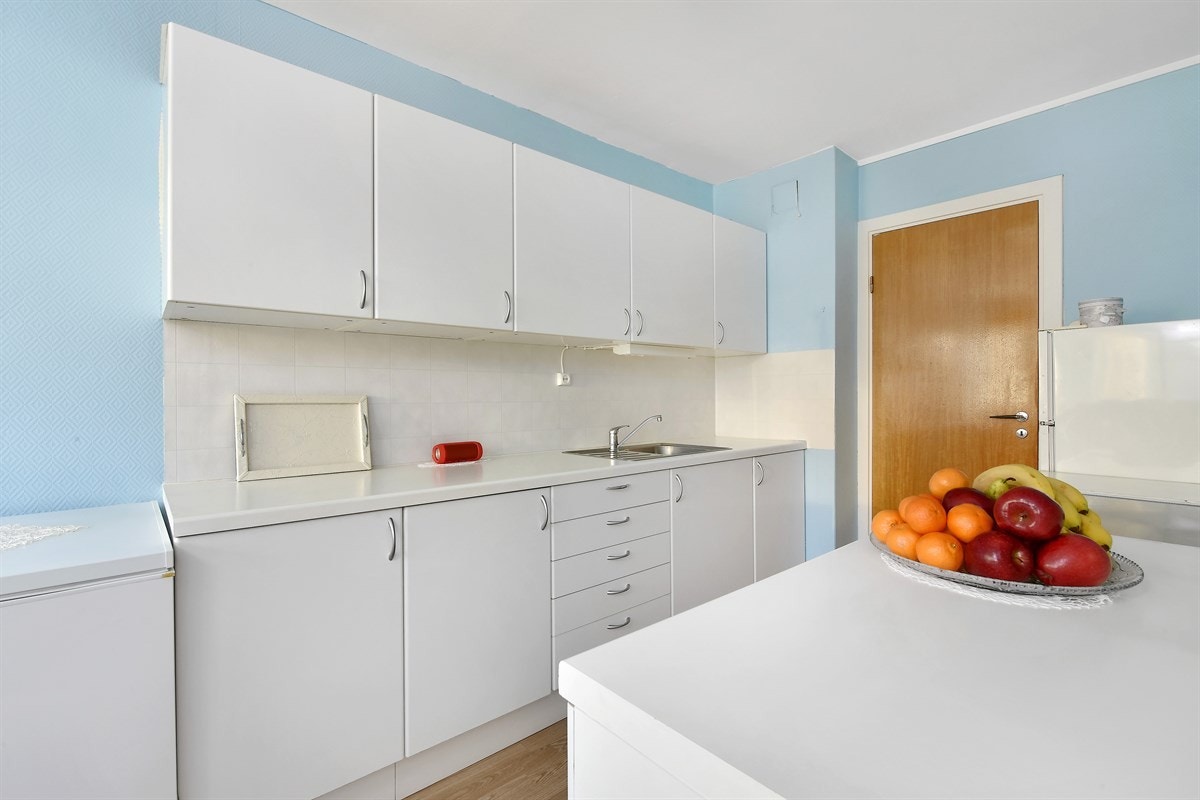 Videre er det laminert benkeplate med enkel vaskekum, stekeovn med koketopp og kjøleskap. Galleribilde