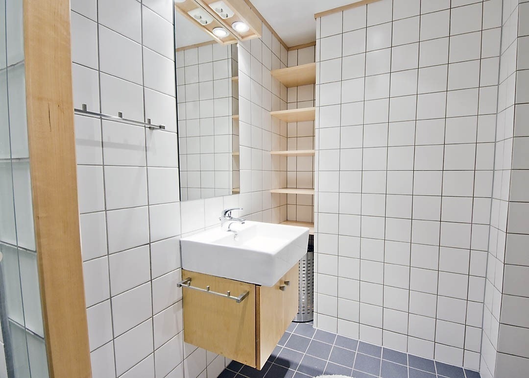 Badet er moderne og elegant med hvite og grå fliser, glassbyggesten og inventar i lyst tre. Dusjhjørne med glassdør, toalett og vask. Det er varmekabler i gulvet og dimbare downlights i taket. Galleribilde