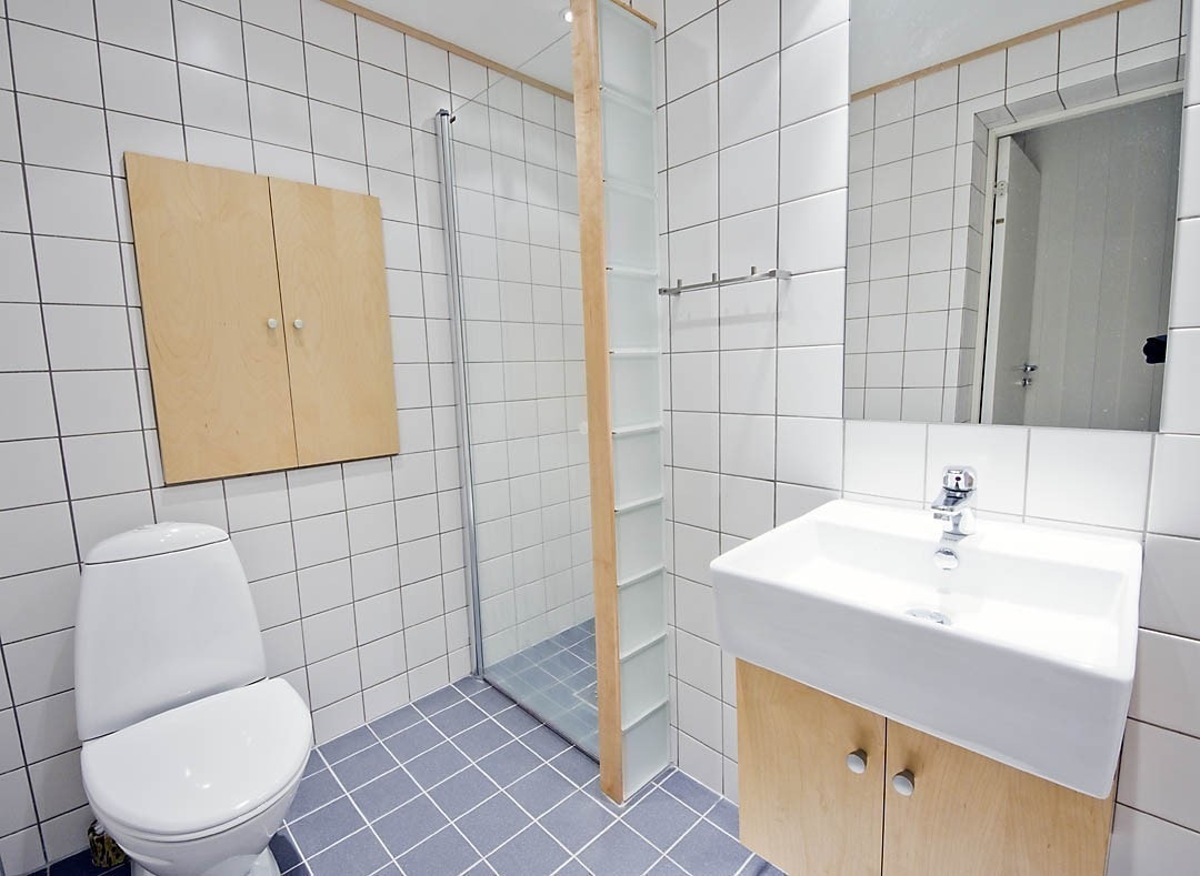 Badet er moderne og elegant med hvite og grå fliser, glassbyggesten og inventar i lyst tre. Dusjhjørne med glassdør, toalett og vask. Det er varmekabler i gulvet og dimbare downlights i taket. Galleribilde