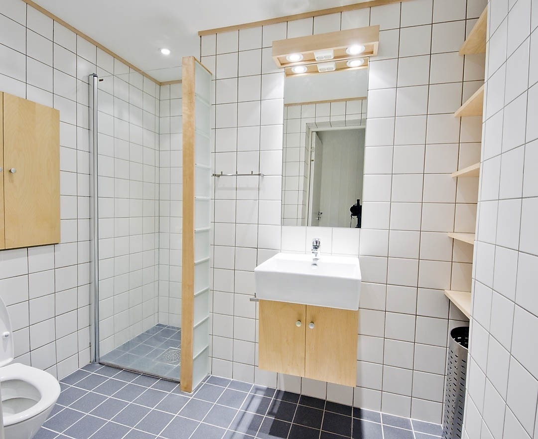 Badet er moderne og elegant med hvite og grå fliser, glassbyggesten og inventar i lyst tre. Dusjhjørne med glassdør, toalett og vask. Det er varmekabler i gulvet og dimbare downlights i taket. Galleribilde