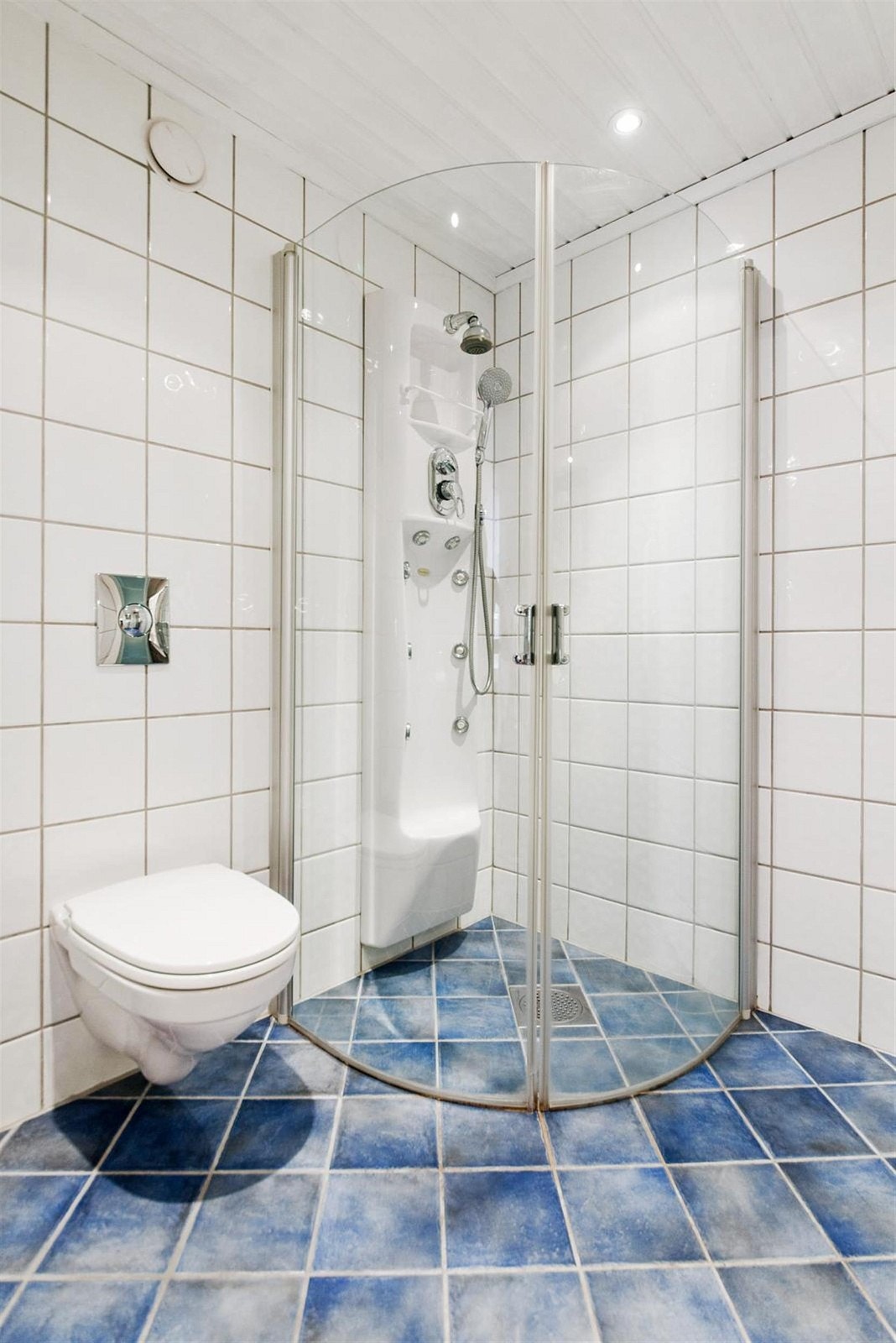 Flislagt bad med veggmontert wc og dusj i hjørne med buede glassdører. Downlights i himling. Galleribilde