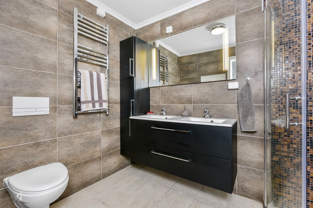 Videre har rommet gulvvarme, elektrisk avtrekksvifte, speil med belysning, heldekkende dobbelservant på servantmøblement, og høyskap. Galleribilde