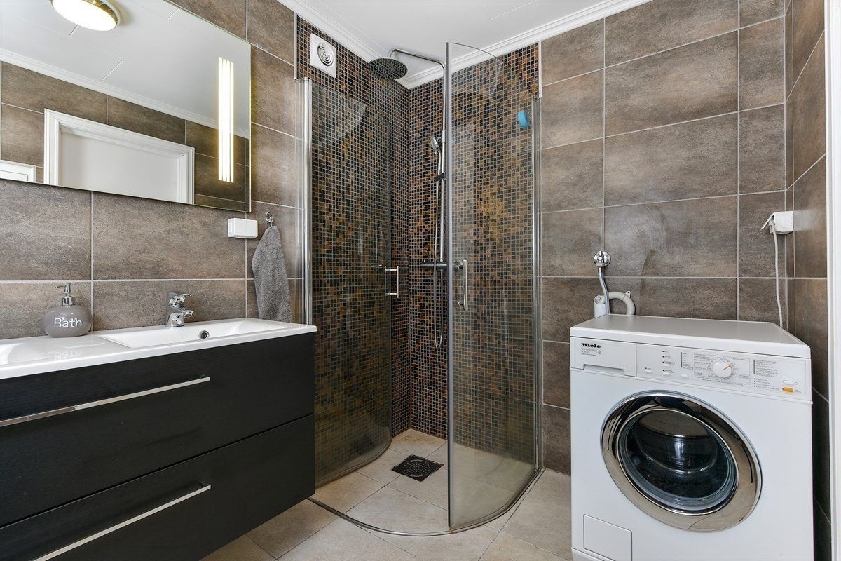 Badet i førsteetasjen er delikat innredet med lysegrå fliser på gulv, fliser i skiftende gråtoner på vegg, samt mosaikkfliser i ulike bruntoner på vegg i dusjsone. Galleribilde