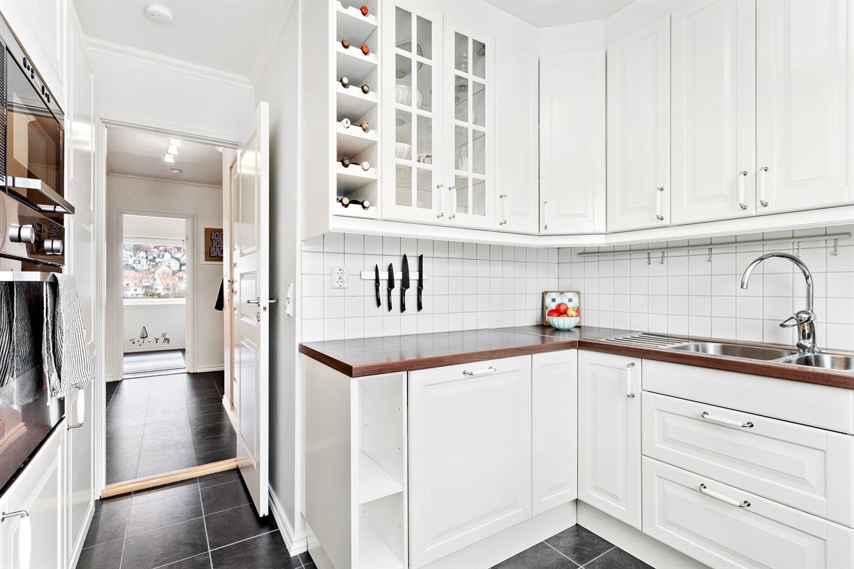 Det er valgt en takhøy kjøkkeninnredning med hvite, profilerte fronter og mørk brun, laminert benkeplate. Mellom over- og underskap er det lagt hvite fliser. Galleribilde