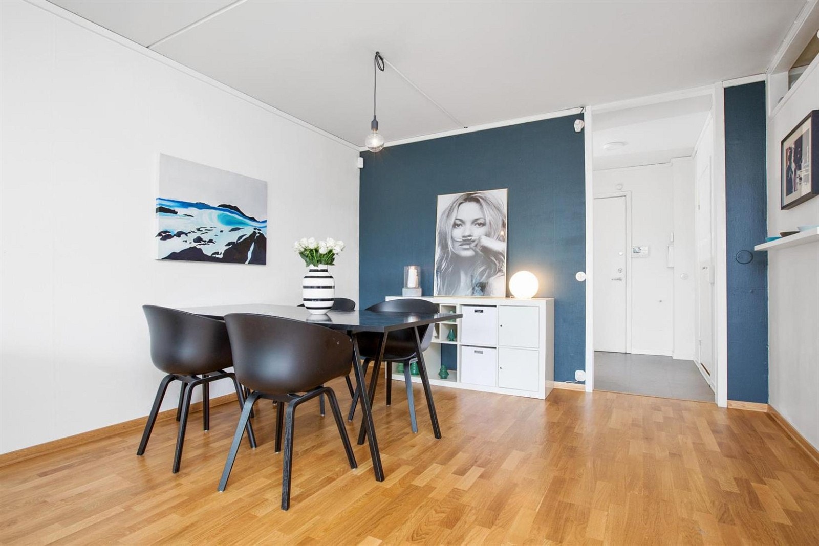 Fin stue med hvite malte vegger samt en kontrastvegg med en tidsriktig blåfarve. Det er inngang fra stuen til delvis åpen kjøkkenløsning. Galleribilde