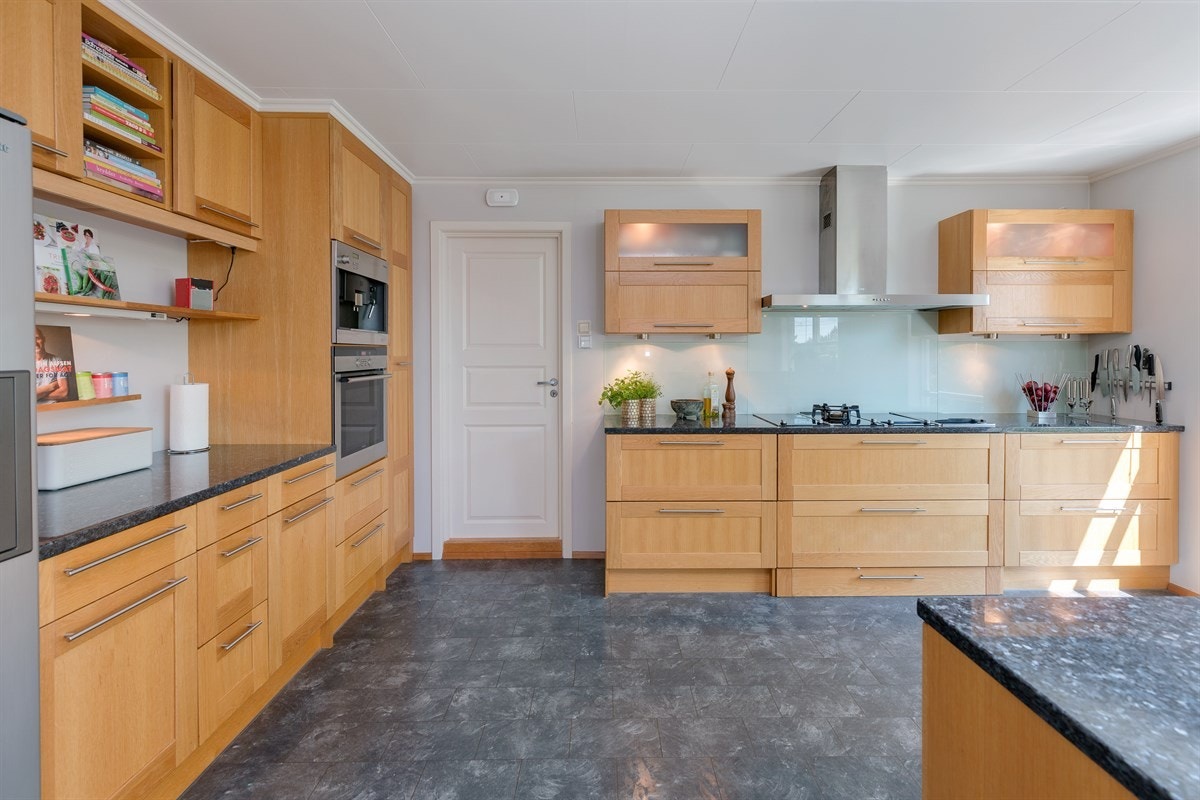 Lyst og innbydende kjøkken med stilrene glatte fronter. Rommet har store vinduer som gir rikelig med lys og bidrar til gode arbeidsforhold ved matlagingen. Galleribilde