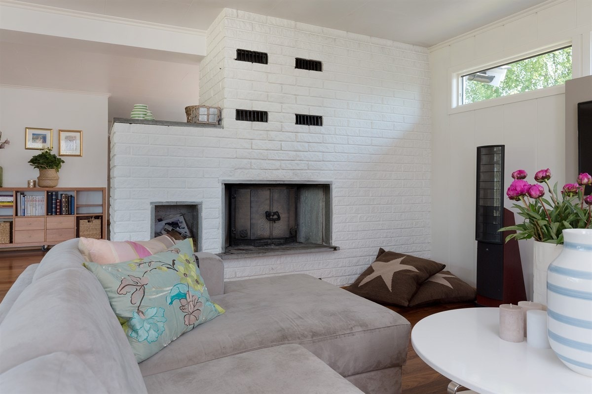 Midt i rommet står en oppmurt peis i malt teglstein, som fungerer som et naturlig skille i denne romslige stuen og avgir en god atmosfære, varme og hygge til rommet. Galleribilde