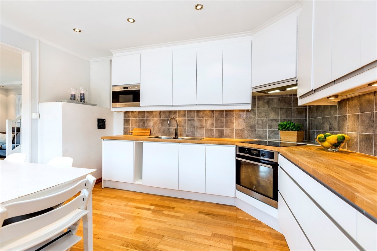 Den takhøye innredningen har hvite, glatte fronter, LED-benkebelysning med dimming, fliser over benk, heltre benkeplate og vaskekum Galleribilde