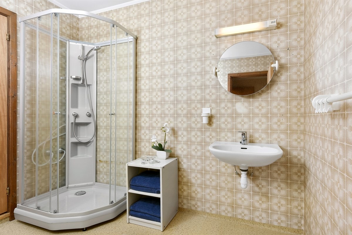 Badet har belegg på gulv og mønstrete baderomstapet på veggene. Videre er det utstyrt med utslagsvask og dusjkabinett. Galleribilde