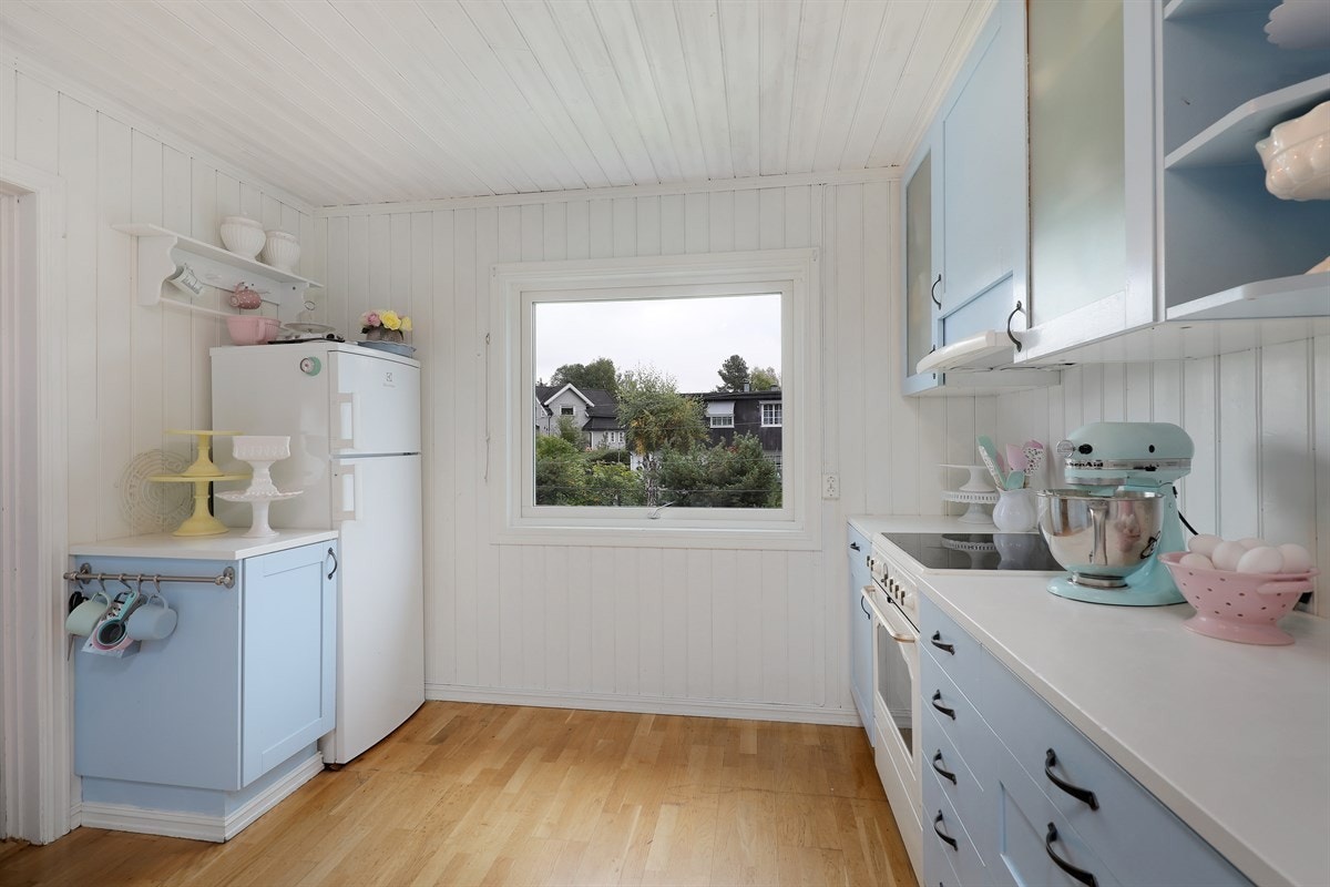 Innredningen består av profilerte fronter med noen vitrinedører, åpne oppbevaringshyller og hvit laminert benkeplate med nedfelt oppvaskkum. Galleribilde