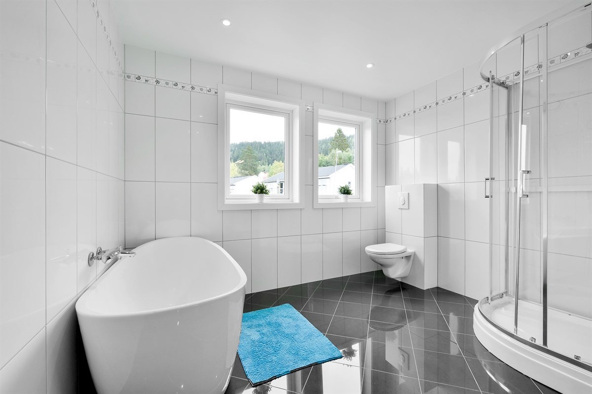 Det romslige badet gir et delikat inntrykk med mørke grå gulvfliser, hvite veggfliser med innslag av dekor, gulvvarme og downlights. Galleribilde