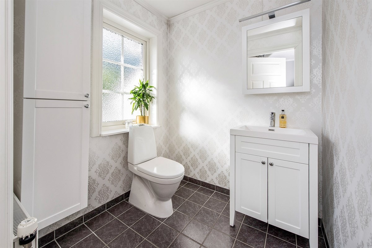 Toalettrommet har gulv med mørke fliser, tapetserte vegger, frittstående toalett og fint lysinnslipp fra vindu. Galleribilde