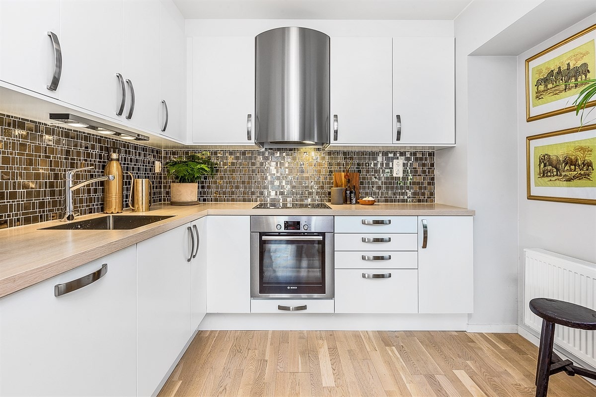 Leiligheten har et tidsmessig og elegant kjøkken designet i en hendig L-form. Innredningen har høye overskap med foring til tak, hvite, glatte fronter, benkebelysning, mørke fliser over benk, og benkeplater i heltre. Galleribilde