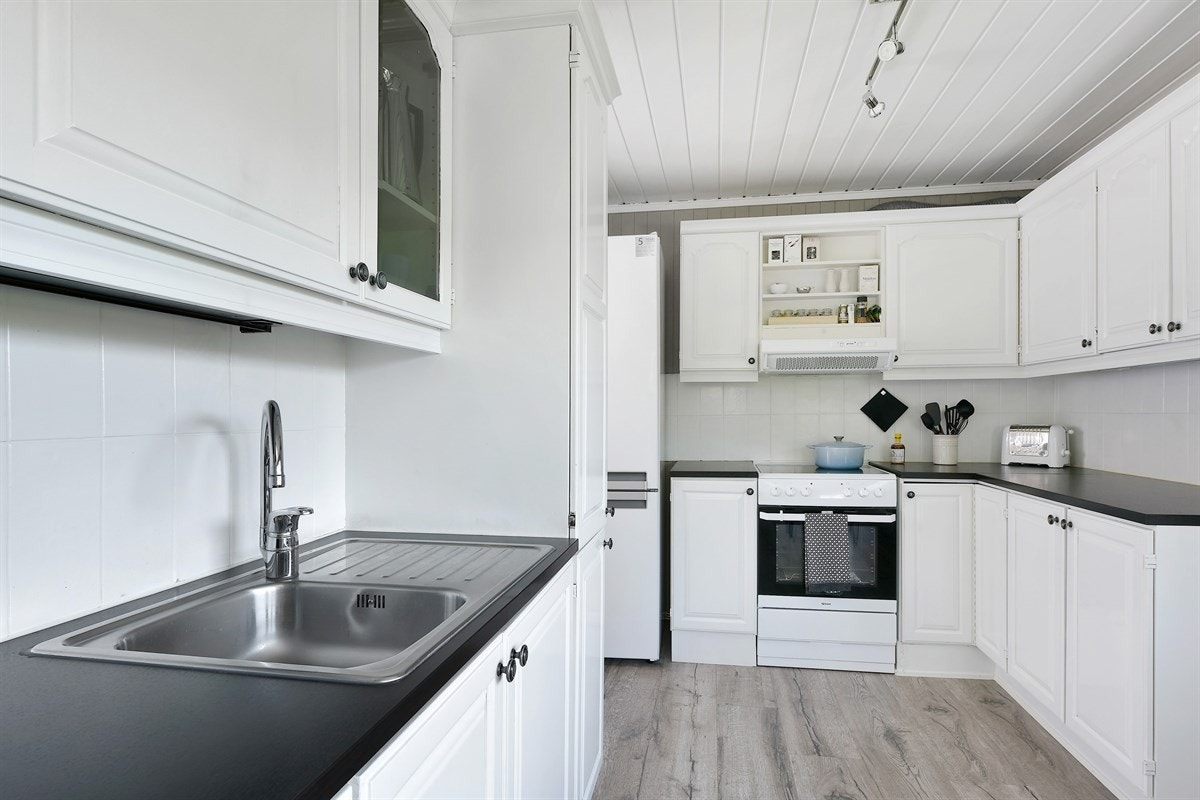 Kjøkkeninnredningen har profilerte fronter, vitrineskap, benkebelysning og fliser over laminert benkeplate med enkel vaskekum. Galleribilde