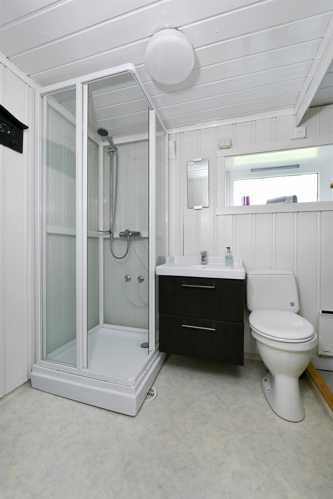 Badet har belegg på gulv, malt trepanel på vegg, heldekkende servant på skuffemøblement, høyskap, gulvmontert toalett og dusjkabinett. Galleribilde