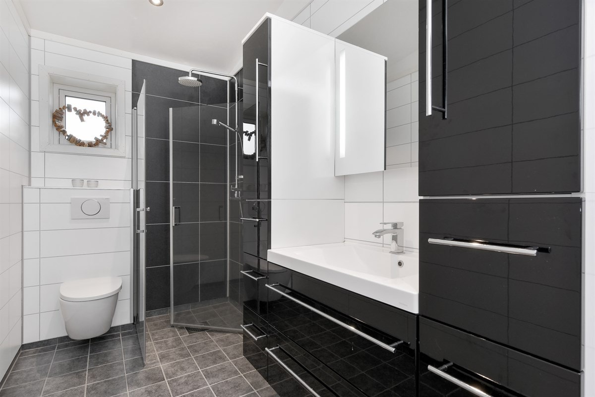 Badet i andre etasje har veggmontert toalett, samt et dusjhjørne med innfellbare glassdører og regnfallsdusj. Innredningen består av et moderne servantmøblement med sorte høyglansfronter, heldekkende servant, to høyskap og speil med belysning. Galleribilde