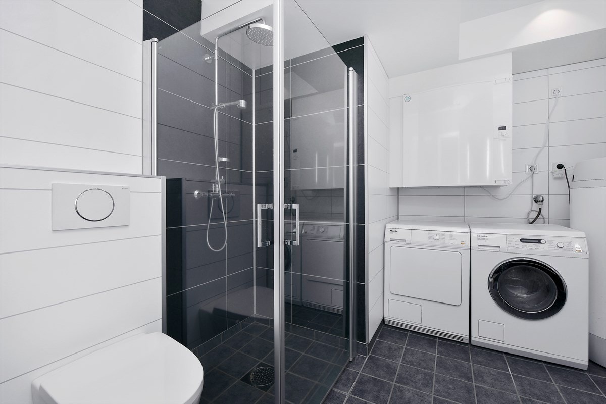 Badet i første etasje er kombinert som vaskerom med opplegg for vaskemaskin og tørketrommel. Videre har badet et veggmontert toalett, dusjhjørne med innfellbare glassdører, regnfallsdusj og mørke vegger i dusjsonen. Galleribilde