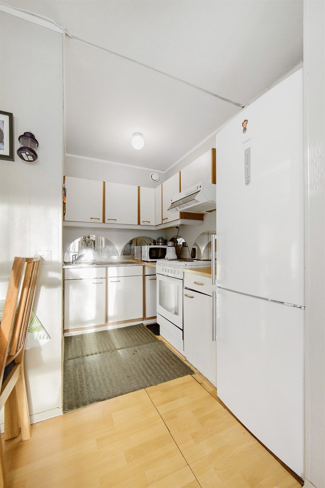 Kjøkkenet har hvite, glatte fronter, buede speilfelt over benk, laminert benkeplate, og heldekkende beslag med enkel oppvaskkum. Galleribilde