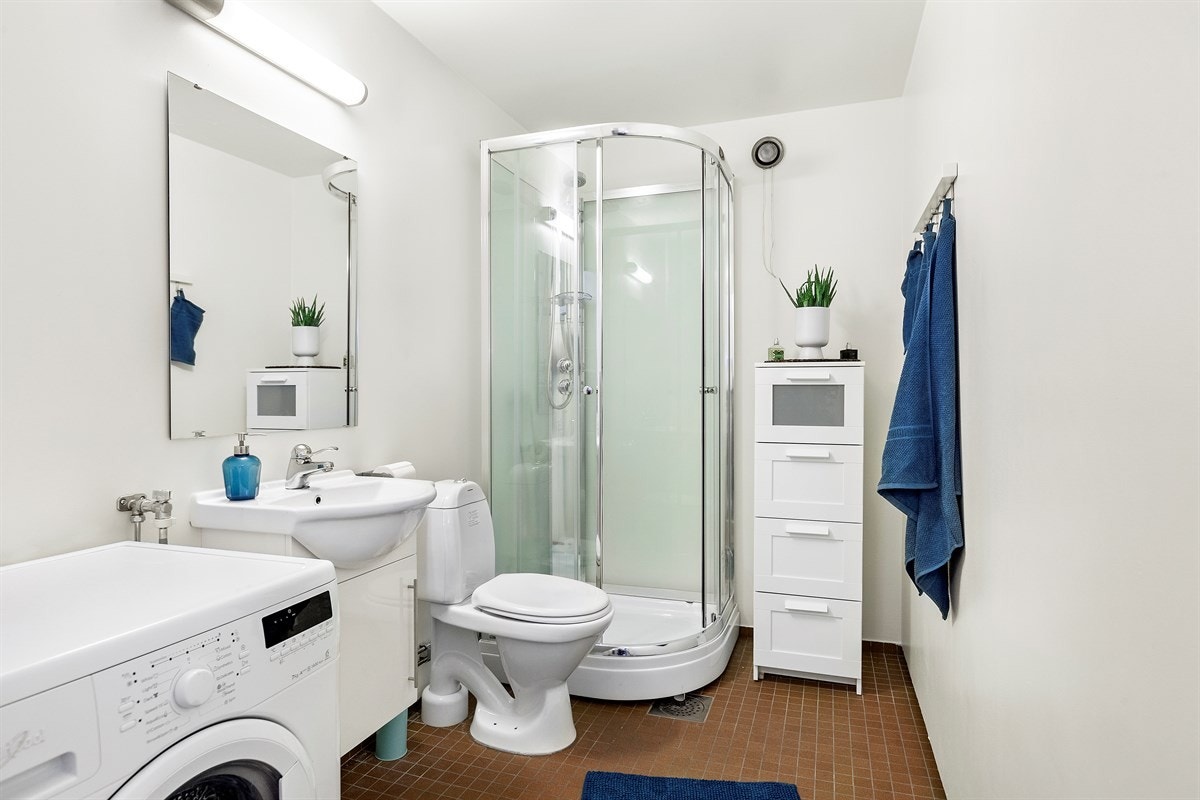 Praktisk baderom med gulvfliser, dusjkabinett, wc, servant og opplegg til vaskemaskin Galleribilde