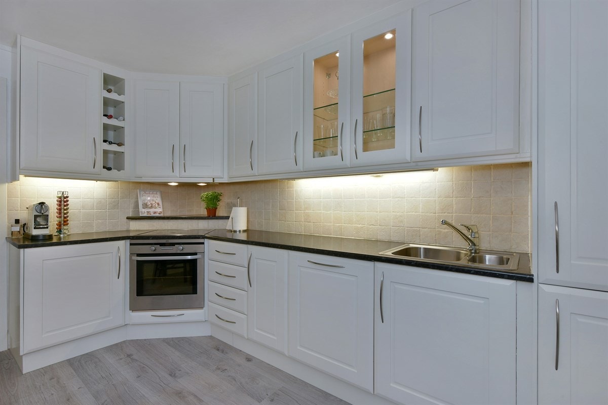 Det takhøye innredningen har hvite, profilerte fronter, vitrineskap med belysning og mørk benkeplate i laminat med dobbel vaskekum. Galleribilde