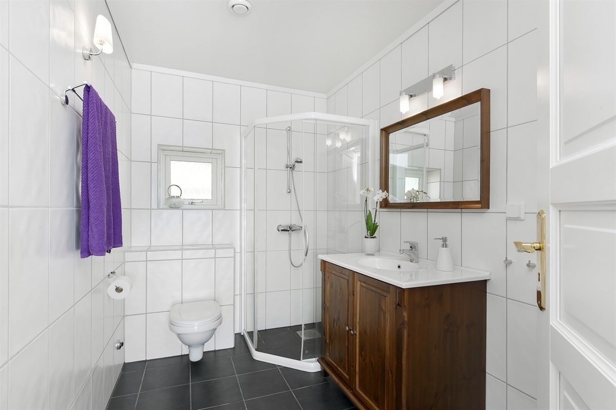 Badet har antrasittgrå fliser på gulv med gulvvarme, hvite veggfliser, heldekkende servant på skap, speil med overlys, veggmontert toalett og dusjhjørne. Galleribilde