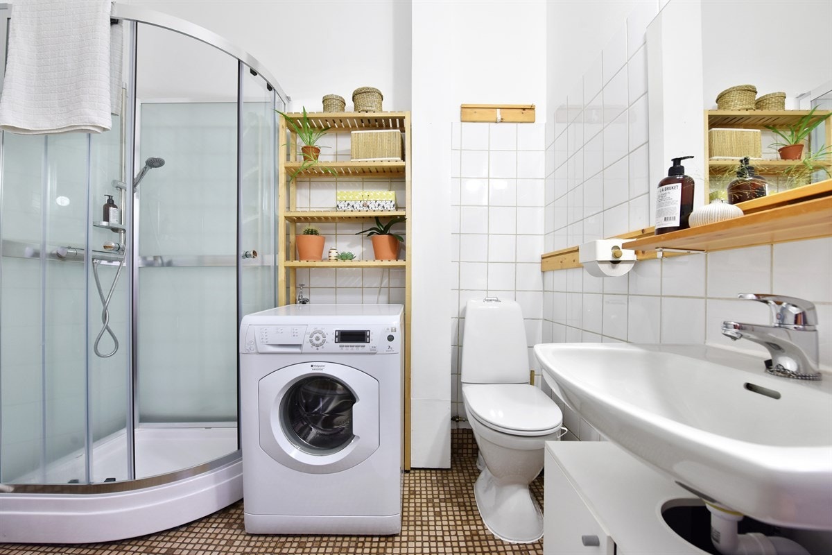 Badet har mosaikkfliser på gulv, lyse fliser på vegger, gulvvarme og opplegg for vaskemaskin og tørketrommel. Galleribilde
