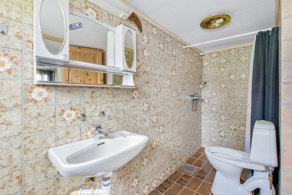 Badet i underetasjen har brune fliser på gulv og lysere fliser på veggene. Innredningen består av enkel servant, speilskap, frittstående toalett og en åpen dusjnisje med stang for dusjforheng. Galleribilde