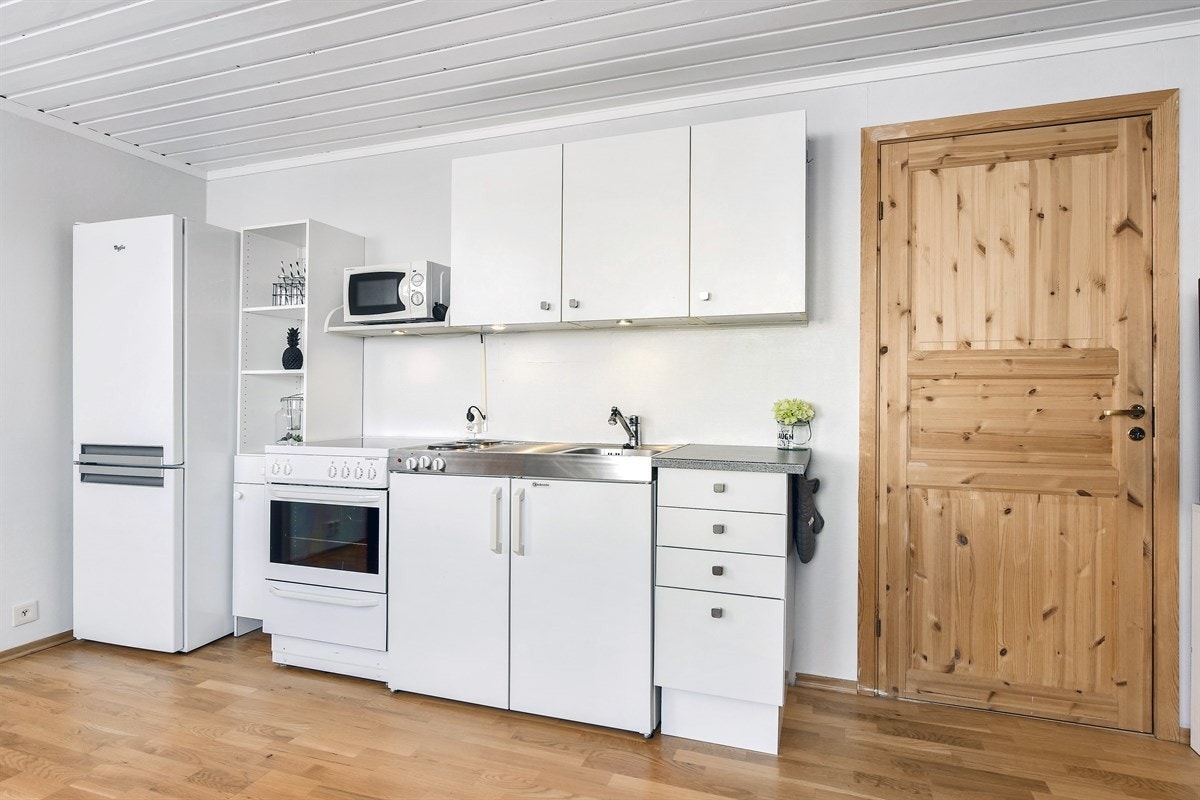 Kjøkkenet har hvite, glatte fronter, en benkeplate i laminat og benkebeslag i stål med nedfelt oppvaskkum og integrert koketopp/kjøleskap. Galleribilde