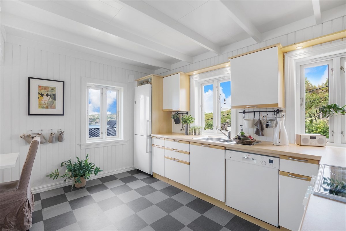 Innredningen er av hvite slette fronter med bøkedetaljer, laminat benkeplate, stålvask med kum, ventilator og fliser over benk ved komfyr Galleribilde