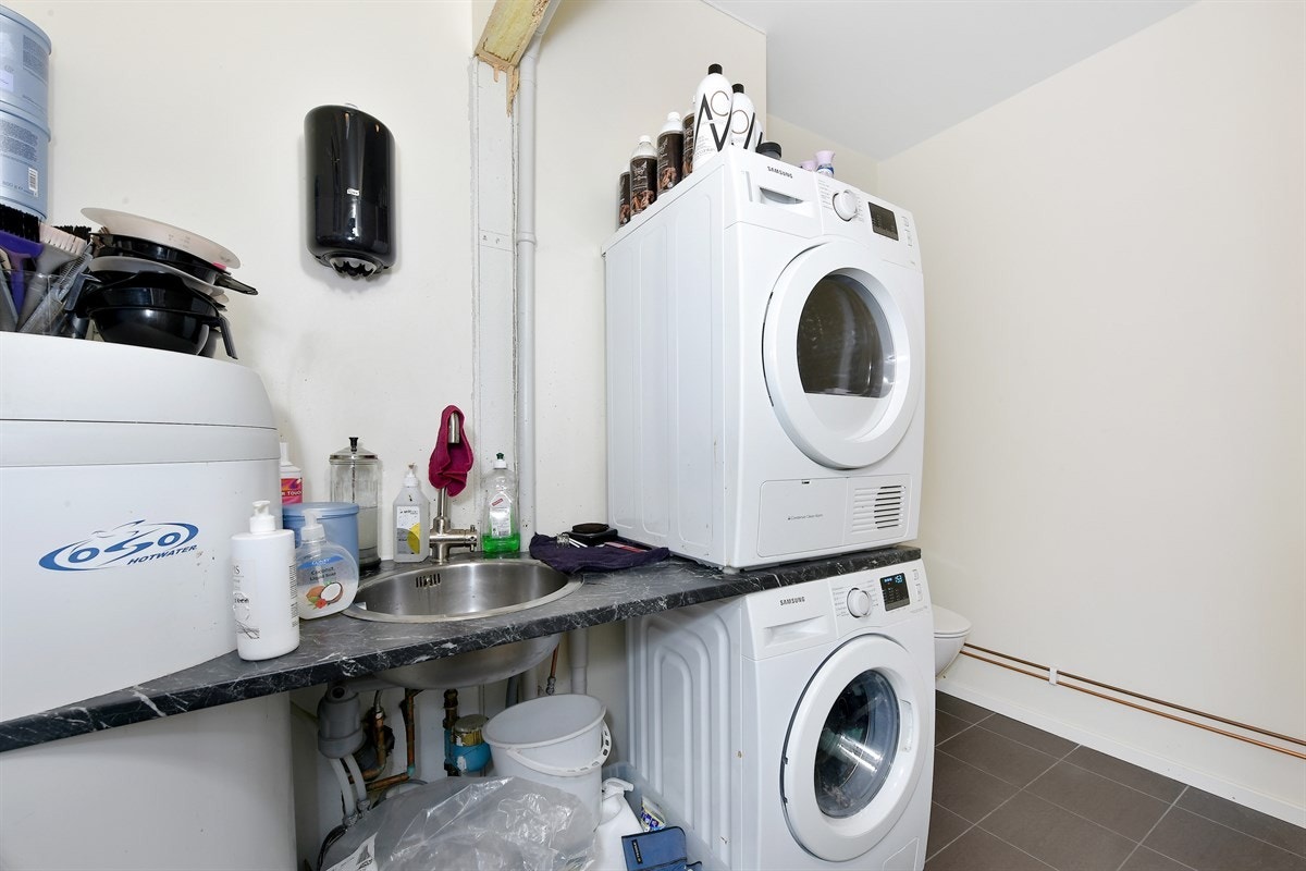 Dette rommet har fliser på gulv, lysmalte veggflater, vaskekum nedfelt i benk, samt opplegg for vaskemaskin, tørketrommel og dusjkabinett. Galleribilde