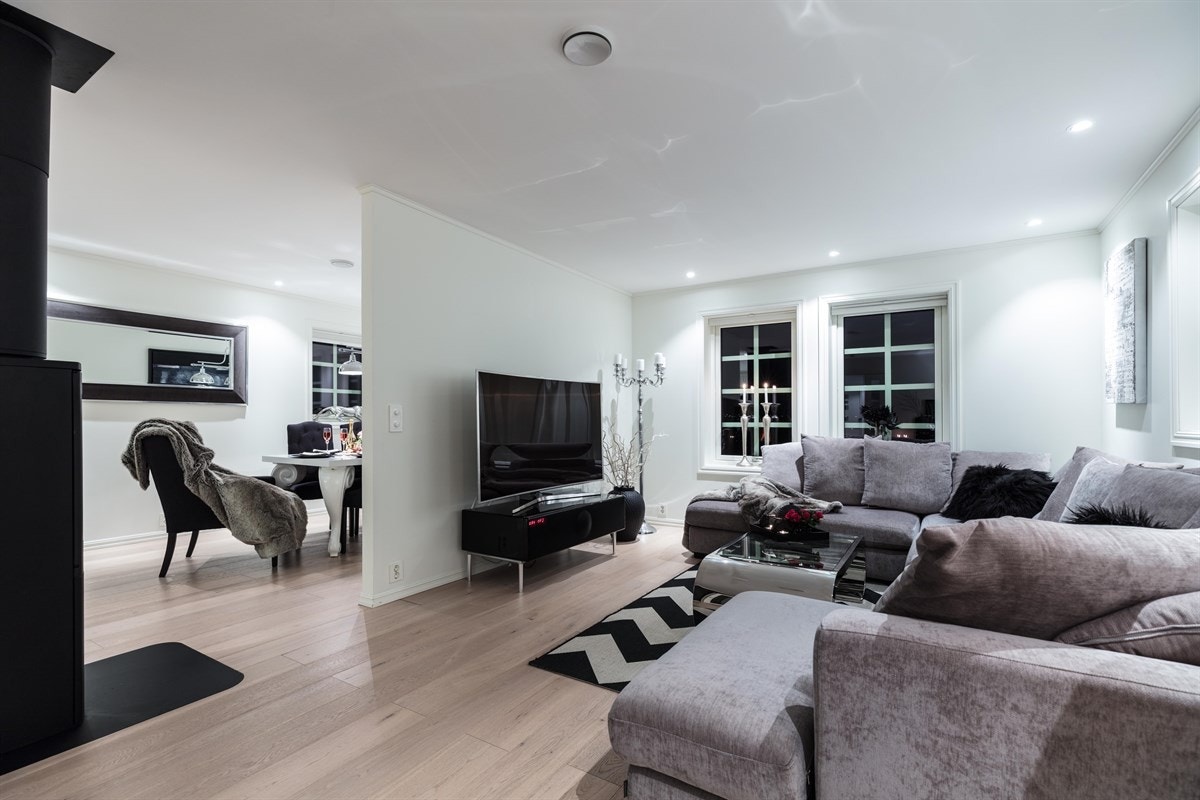 En mørk fondvegg bidrar med karakter og innfelte downlights i himling skaper ytterligere stemning. Stuen har tofløyet dør til terrasse, og i overgangen til spisestuen er det satt inn en moderne peisovn for ekstra varme og hygge. Galleribilde