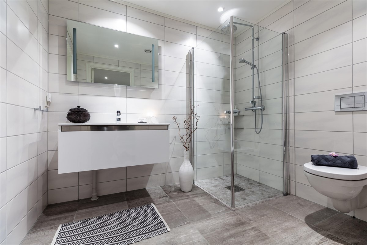 Badet i første etasje er tilknyttet entreen, og har flislagte overflater, integrerte downlights i himling og varme i gulvet. Rommet har speil med integrert belysning, heldekkende servant på møblement, vegghengt toalett og et dusjhjørne med regnfallsdusj. Galleribilde