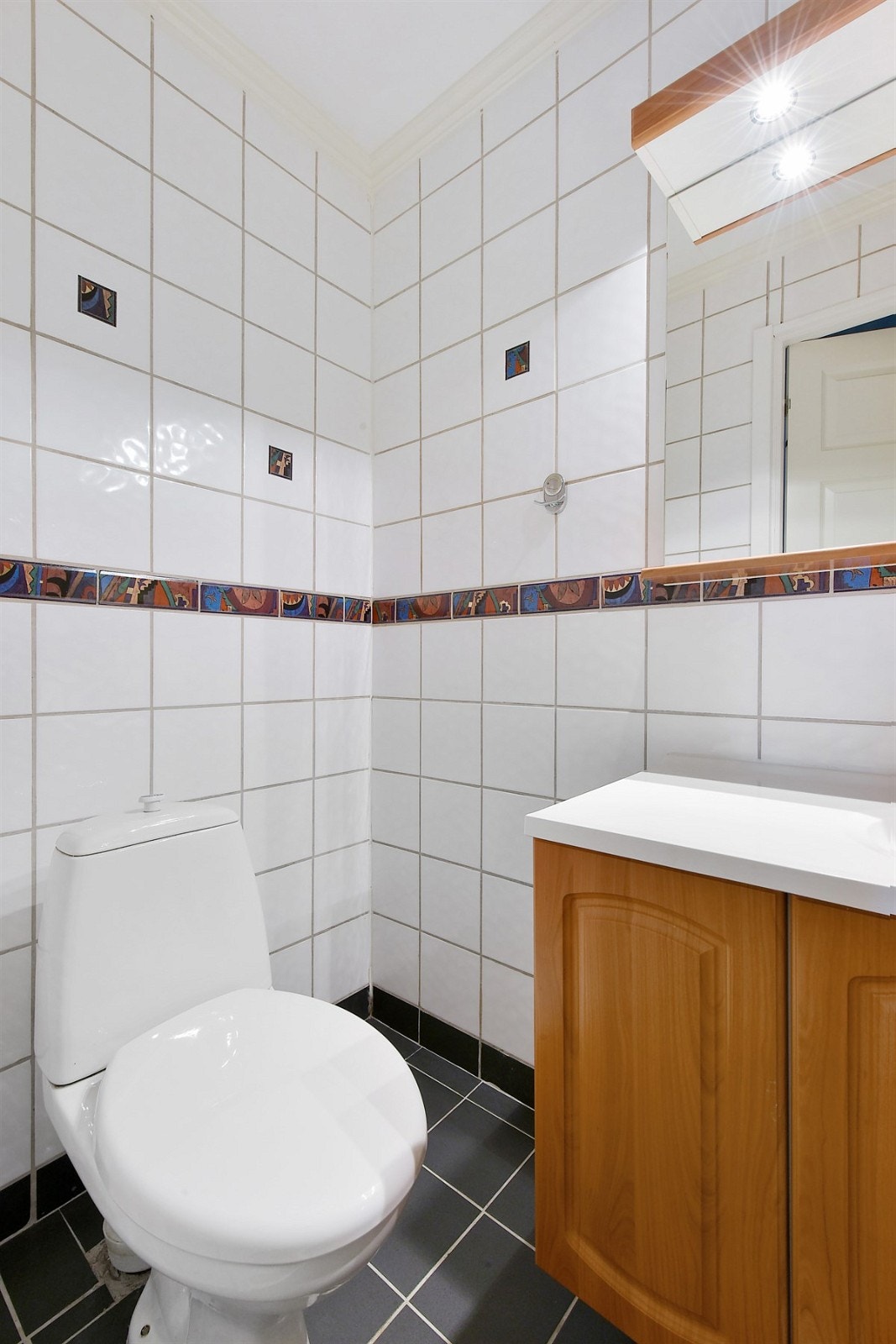 Badet er utstyrt med dusjhjørne med skyvedører, frittstående toalett og heldekkende servant med tilhørende skap og hyllemøblement, samt speil med belysning. Galleribilde