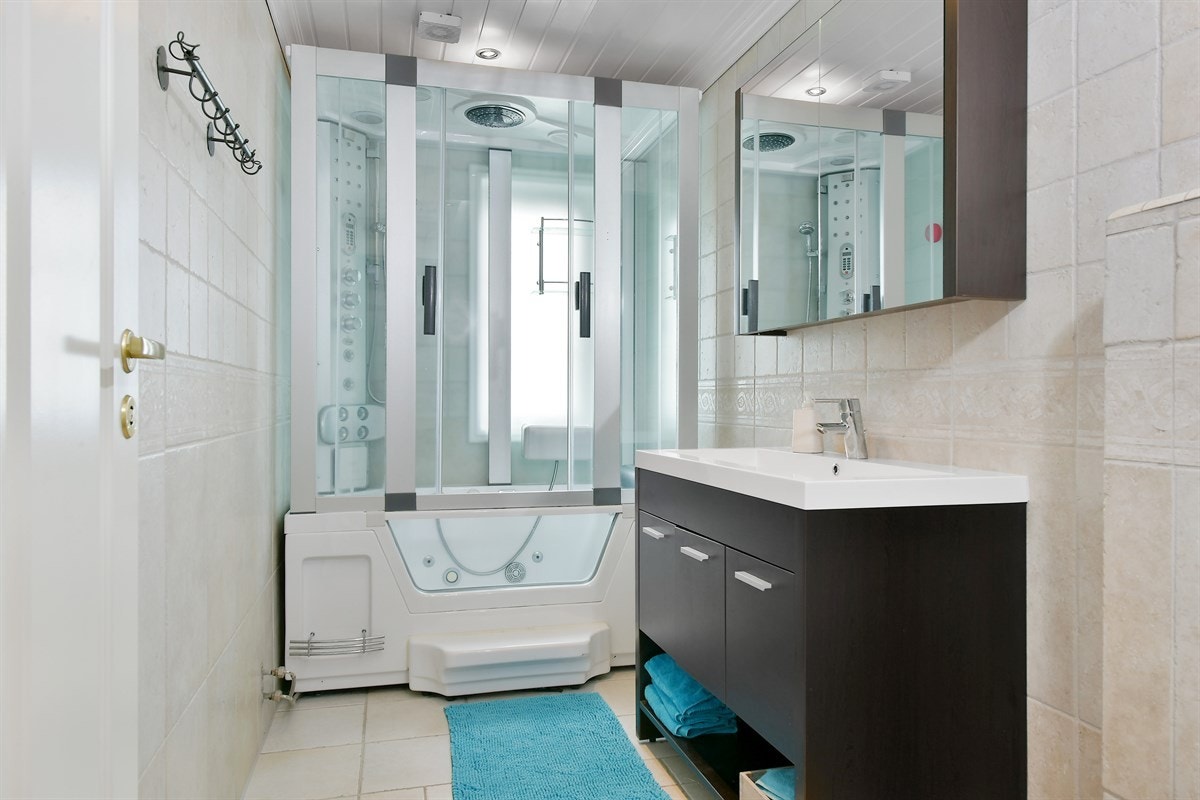 Rommet er utstyrt med fliser på vegg og gulv, gulvvarme, elektrisk avtrekksvifte og veggmontert toalett. Videre er det steamdusjkabinett med boblebad og heldekkende servant med romslig skapmøblement og skapspeil. Galleribilde