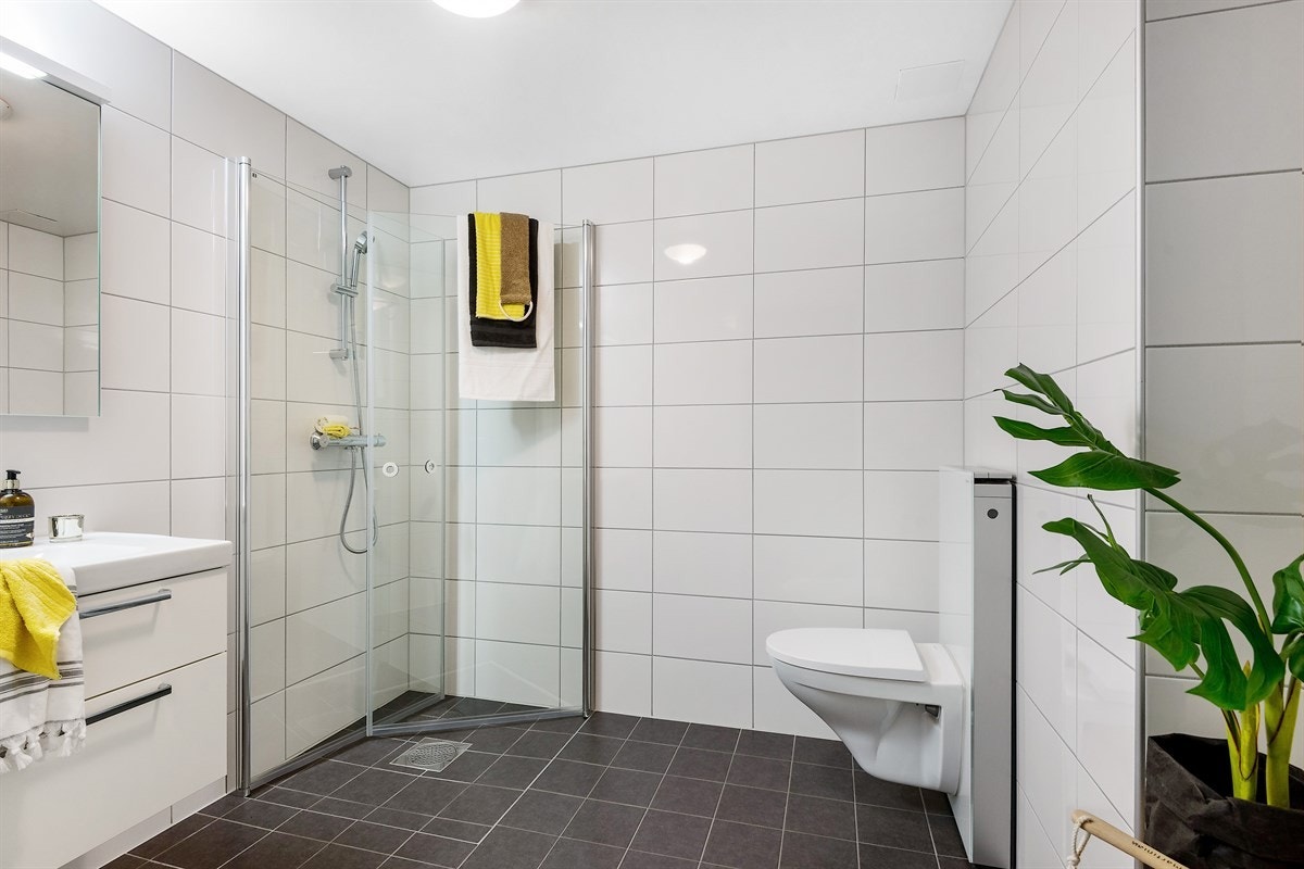 Meget pent flislagt bad med romslig dushjørne med pen dusjgarnityr på vegg med stilrene, innfellbare dusjevvegger i herdet glass. Vegghengt klosett. Galleribilde