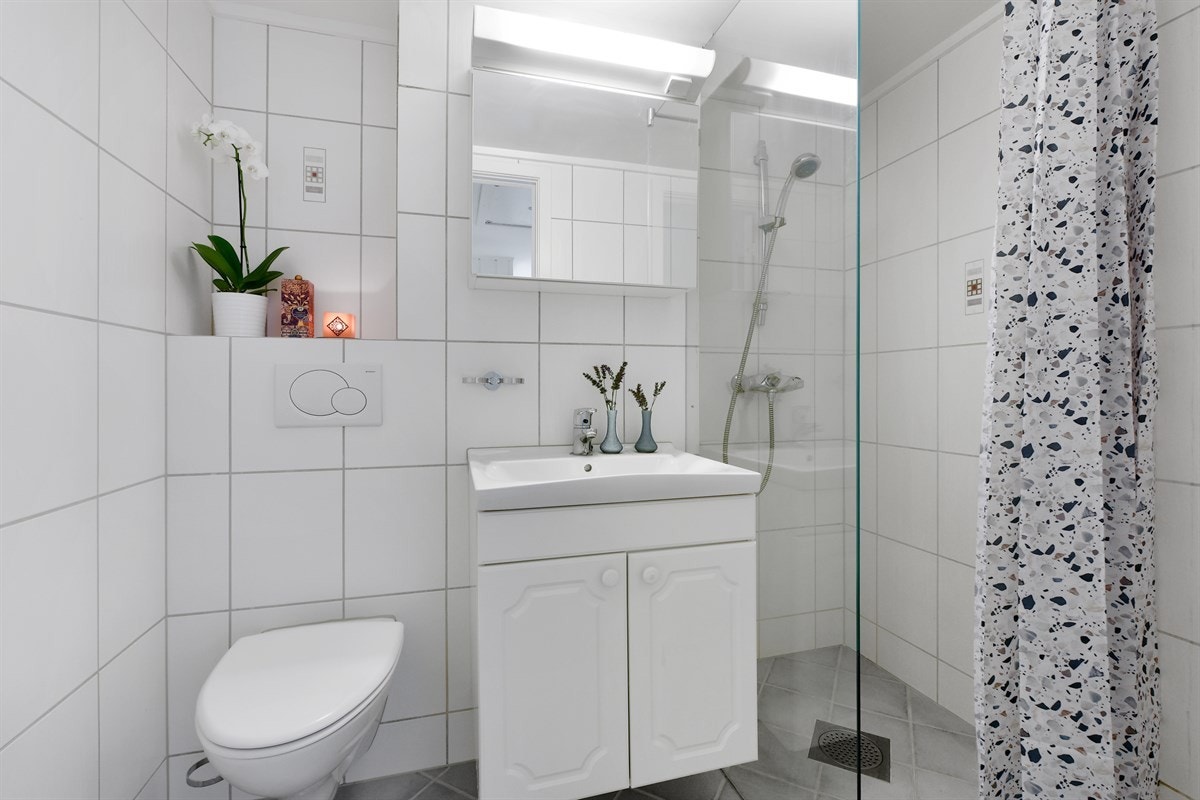 Badet er delikat med grå gulvfliser og hvite veggfliser, samt downlights og gulvvarme. Badet har vegghengt toalett og et nytt dusjhjørne med vegg i herdet glass fra 2017/18. Galleribilde