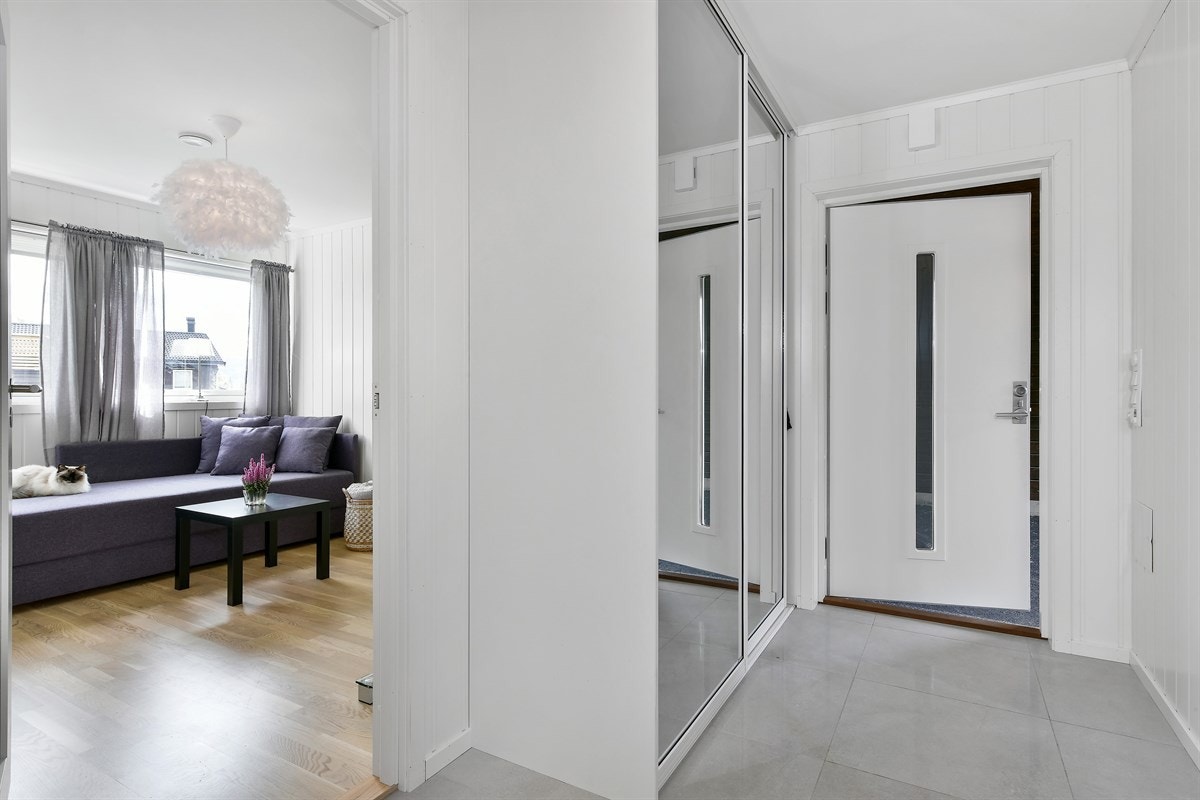 Boligen har trappefri adkomst via carport inn til en pen entré. Rommet har store, lysegrå fliser på gulv og hvitmalte panelplater på vegg. Galleribilde