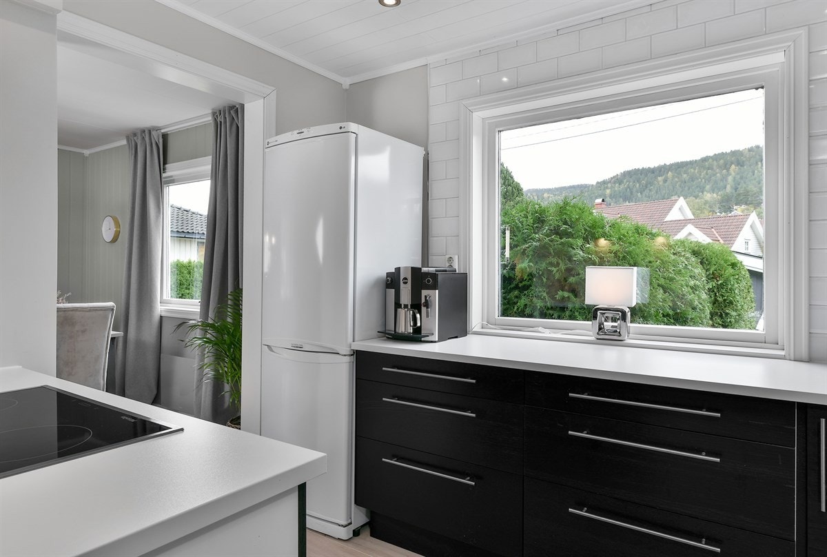 Kjøkkenet er stilrent og praktisk designet med takhøye overskap, gode benkeflater og rene linjer. Kjøkkeninnredningen har sorte, finerte fronter og en hvit laminatbenkeplate med nedfelt kum i stål. Galleribilde