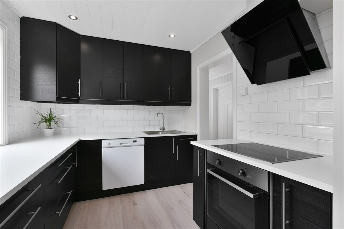 Videre er kjøkkenet utstyrt med integrerte hvitevarer som koketopp, stekeovn og oppvaskmaskin, samt et frittstående kjøleskap. Rommet har hvite fliser på veggene, downlights i himling og en sort, moderne ventilator som står godt til resten av interiøret. Galleribilde