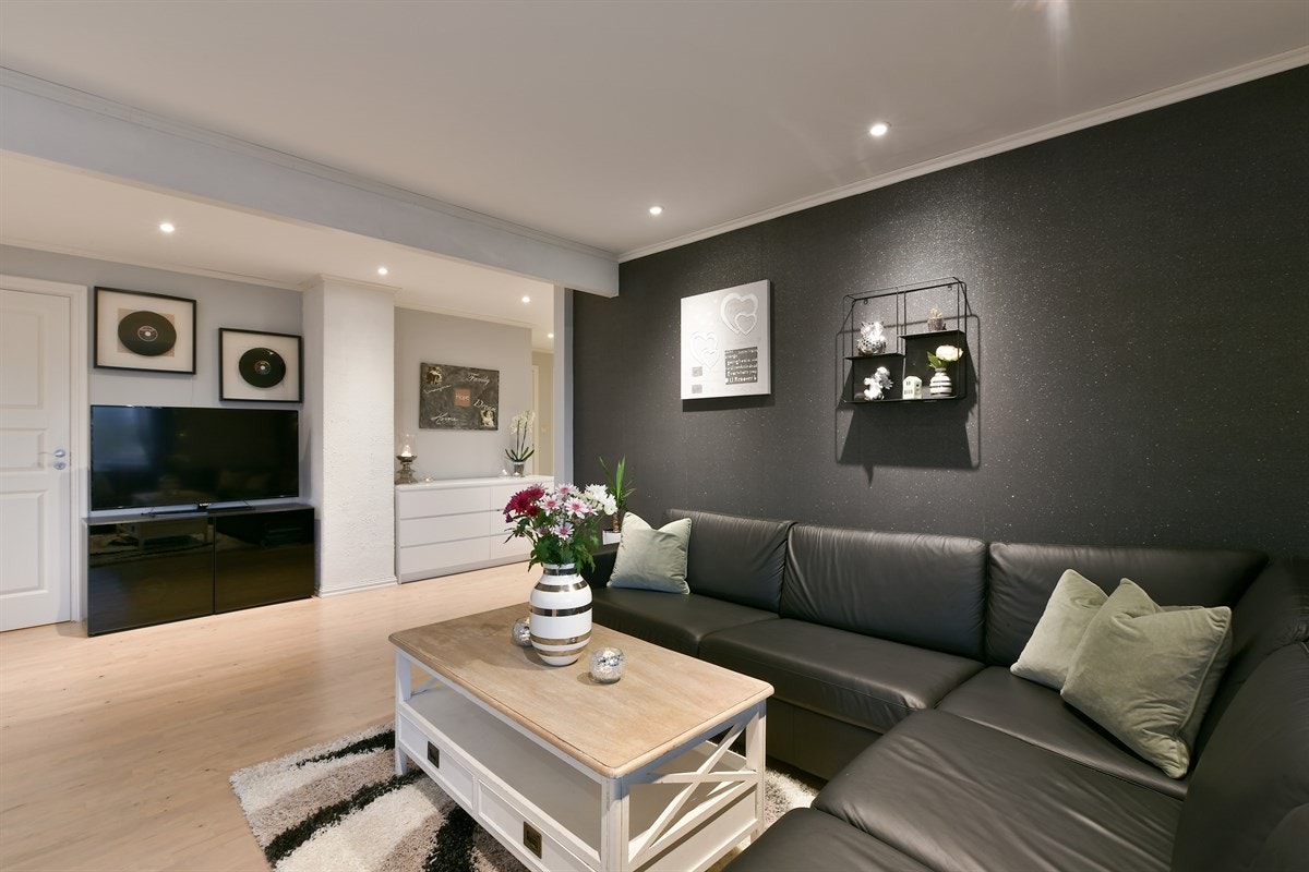 Videre har stuen lyse vegger og én vegg i en moderne, mørk kontrastfarge. Gulvet har parkett og gulvvarme, og i himlingen er det downlights. Galleribilde