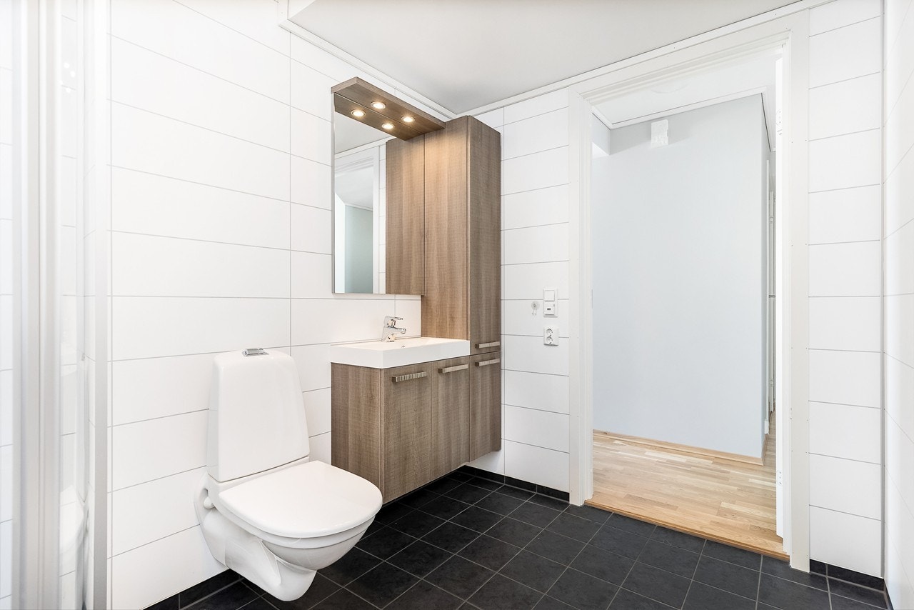 Badet er utstyrt med veggmontert toalett, dusjhjørne og heldekkende servant på en lekker skapseksjon. Her har du rikelig med oppbevaringsplass Galleribilde